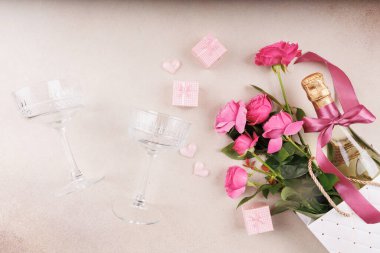 Şampanya şişesi, pembe güller, küçük hediyeler ve tarafsız dokulu arka planda iki zarif bardak..
