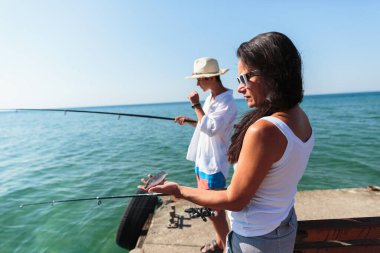 A teenage boy and his mother fish together at a seaside pier, with the mother holding a small fish and the boy managing his fishing rod under a clear sunny sky clipart