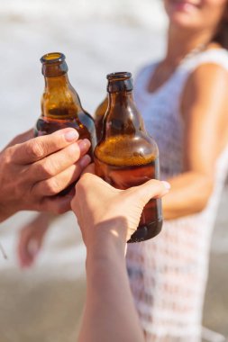 Arkadaşların kadeh kaldırarak bira şişelerini kaldırdığı beyaz örgü ören sahil kıyafetli gülümseyen bir kadın ve arka planda bulanık okyanus dalgaları..