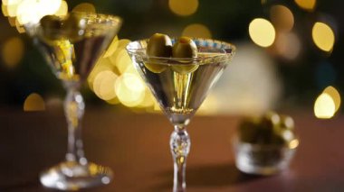 Bir kadın eli zarif bir şekilde zeytinli bir martini bardağı kaldırır, sıcak bokeh ışıkları ile neşeli bir ortam yaratır. Yüksek kaliteli FullHD görüntüler