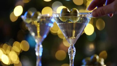 Bir kadın, arka planda zarif bokeh ışıkları parlayan bir martini bardağından zeytinleri nazikçe kaldırır. Yüksek kaliteli FullHD görüntüler
