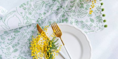 Çiçek desenli bir peçetede altın çatal bıçak, beyaz bir tabak ve mimoza çiçekleri bulunan zarif bir bahar sofrası..
