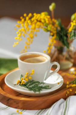 Tahta tepside bir fincan kahvenin bulunduğu narin bir bahar sahnesi, canlı mimoza çiçekleri ile sıcak atmosferi güçlendiriyor..
