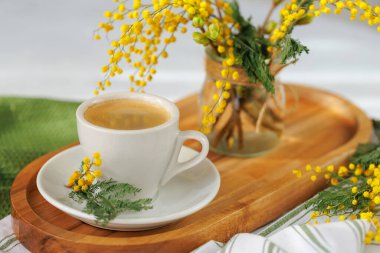 Beyaz bir fincan kahve ahşap tepside mimoza çiçekleriyle, huzurlu ve ılık bir bahar sabahını çağrıştırıyor..