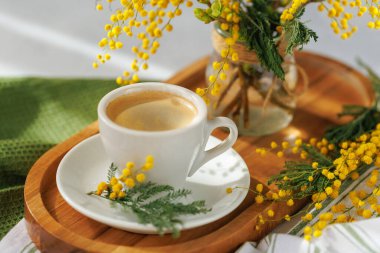 Bir fincan taze demlenmiş kahve, narin mimoza çiçekleriyle çevrili ahşap bir tepside oturur. Rahatlatıcı bir bahar anı için mükemmeldir..