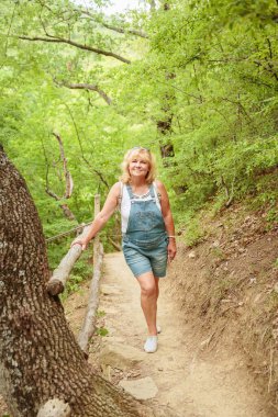 Orta yaşlı şort giymiş bir kadın sırt çantasıyla orman yolunda yürüyor. Çekici kadın gezgin yürüyüş parkurunda rahatlıyor ve doğanın tadını çıkarıyor. Seyahat ve sağlıklı yaşam tarzı