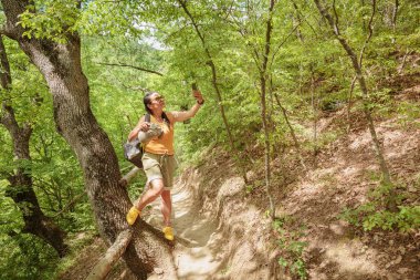 Orman yolunda yürüyen ve akıllı telefon kullanan bir kadın. Çekici kadın gezgin yürüyüş yolunda rahatlıyor ve doğanın tadını çıkarıyor.