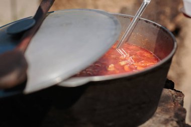 Büyük metal bir tencere kısmen kaplı, sebze ve et parçalarıyla dolu kırmızı bir yahni ile kaynayan, açık bir ateşte, kırsal ve doyurucu bir pişirme sahnesi yaratıyor doğal bir ortamda.