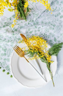 Altın çatal bıçak, beyaz tabak ve sarı mimoza çiçekleri desenli bir peçeteye yerleştirilmiş zarif bir bahar esinlenilmiş masa takımı..