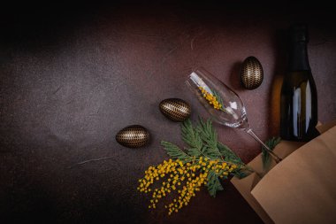 Bir şişe şampanya, içinde mimoza çiçekleri olan bir bardak ve şenlik havası için desenli bir yüzeye yerleştirilmiş altın yumurtalar..