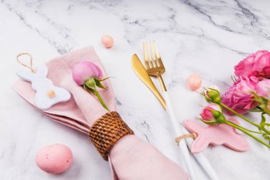 Pembe peçeteli Paskalya masası, keçeli tavşan süsü, altın çatal bıçak, benekli yumurta ve mermer arka planda taze pembe güller.