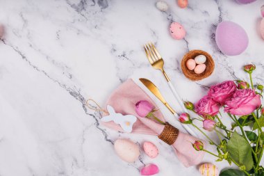 Paskalya temalı masa dekorunun üst görüntüsü pembe güller, pastel yumurtalar, altın kaplar ve tavşan peçetesi halkası.