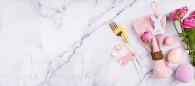 Pembe peçeteli yatay düzen, altın kaplar, tavşan süsleri, Paskalya yumurtaları ve beyaz mermer üzerinde çiçekler.