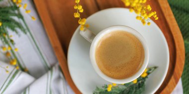 Tahta bir tepside beyaz bir kahve fincanının yukarıdan görünüşü, mimoza çiçekleriyle güzelce düzenlenmiş, mükemmel bir bahar kahvaltısı sahnesi yaratıyor..