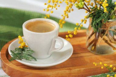 Sıcak espresso ile beyaz bir seramik fincan ahşap bir tepside duruyor, taze mimoza çiçekleriyle süslenmiş, huzurlu bir bahar sabahının özünü yakalıyor..