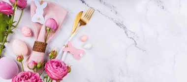 Beyaz tavşan süslemeli Paskalya peçetesi, altın çatal bıçak, mermer yüzeyde pastel yumurta ve çiçekler.