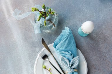 Yumurta tutacağı, çiçek açan vazo, beyaz tabak ve çatal bıçaklı Paskalya masasının en üst görüntüsü.