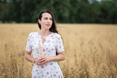 A beautiful brunette woman in a white dress runs along a field of golden chickpeas ear. Stylish girl in the field in windy weather