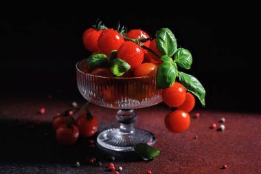 Fesleğen yapraklı kırmızı kiraz domatesleri yumuşak gölgeli karanlık bir yüzeyde zarif bir cam kasede.