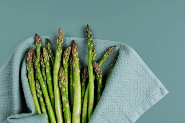 Yumuşak mavi arka planda dokulu mutfak havlusuna sarılı bir demet yıkanmış yeşil kuşkonmaz.