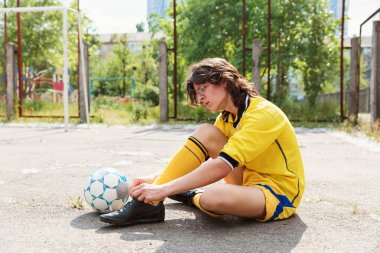 Sarı futbol formalı genç asfaltta oturuyor ve antrenmandan önce ayakkabı bağcıklarını bağlıyor.