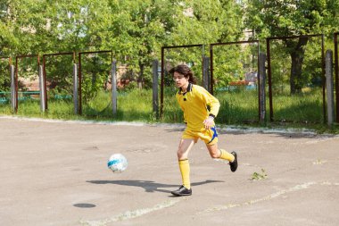 Sarı spor üniformalı genç, dışarıda asfalt sahada futbol topunun peşinde koşuyor.