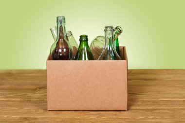 Ahşap masanın üzerine yeşil arka planda çeşitli cam şişeler yerleştirilmiş karton kutu.