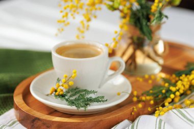 Sıcak bir bahar sabahı sahnesi, ahşap tepside beyaz bir kahve fincanı, taze mimoza çiçekleriyle süslenmiş, sıcaklığı ve cazibesi yayılmış..