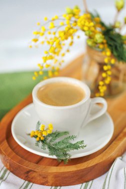 Sıcak bir bahar sabahı sahnesi, ahşap tepside bir fincan kahve, taze mimoza çiçekleriyle süslenmiş, sıcak ve neşeli bir hava yaratıyor..