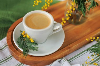 Bir fincan taze demlenmiş kahve ahşap tepside, sarı mimoza çiçekleriyle süslenmiş, parlak ve samimi bir bahar havası yaratıyor..