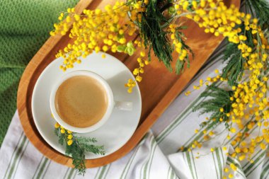 Bir fincan taze demlenmiş kahve ahşap bir tepside, arka planda mimoza çiçekleri vazosu, rahat bir bahar hissi yaratır..