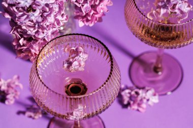 İçi köpüklü şarap ve mor arka planda büyük bir buketin yanında süzülen leylak çiçekleriyle dolu iki şampanya bardağının yukarıdan görünüşü