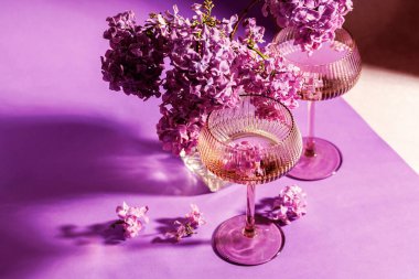 Işıl ışıl şarap kadehleri ve eflatun demeti, koyu gölgeler ve çiçek desenli menekşe bir yüzeye dizilmiştir.