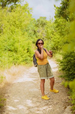Orta yaşlı şort giymiş bir kadın sırt çantasıyla orman yolunda yürüyor. Çekici kadın gezgin yürüyüş parkurunda rahatlıyor ve doğanın tadını çıkarıyor. Seyahat ve sağlıklı yaşam tarzı