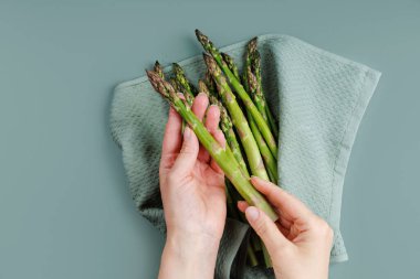 Doğal yemek hazırlığı için mutfak havlusunun üzerinde temiz yeşil kuşkonmaz mızrakları tutan kadın elleri.