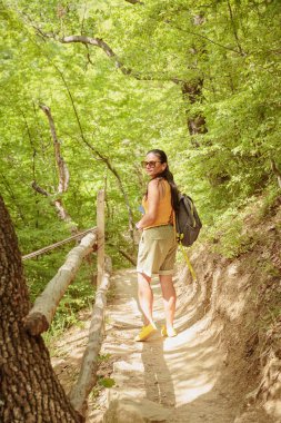 Orta yaşlı şort giymiş bir kadın sırt çantasıyla orman yolunda yürüyor. Çekici kadın gezgin yürüyüş parkurunda rahatlıyor ve doğanın tadını çıkarıyor. Seyahat ve sağlıklı yaşam tarzı