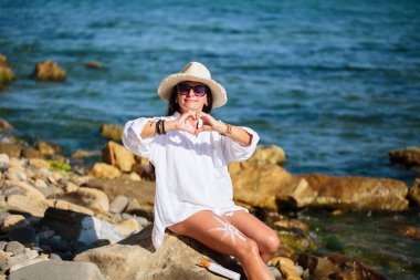 Gülümseyen kadın ellerle kalp yapıyor. Kıyı kayalıklarında otururken, bacağında güneş kremi ile.