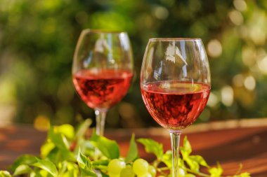 Taze üzümlü iki bardak gül şarabı ve bahçenin arka planındaki ahşap yüzeyde yeşil yapraklar.