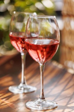 Ahşap bir masada iki gül şarap kadehi. Arkaplanda canlı yansımalar ve bahçe örtüsü var.