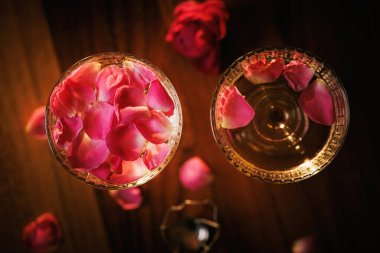 İki kristal şampanya kadehi, süzülen gül yaprakları ve ahşap bir yüzey üzerinde ışıldayan mum ışığında romantik bir akşam..