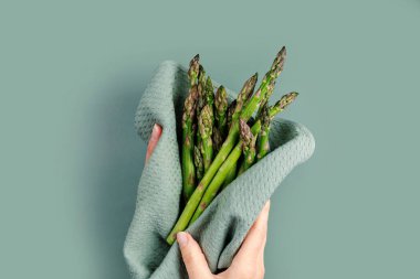 Kadın eli, pastel bir yüzeyde mutfak havlusuna sarılı taze kuşkonmaz paketini tutuyor.