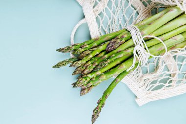 Yeşil kuşkonmaz demetinin üst görüntüsü yeniden kullanılabilir bir ağ torbasının içine sarılmış olarak pastel mavi arka planda fotokopi alanı ile birlikte