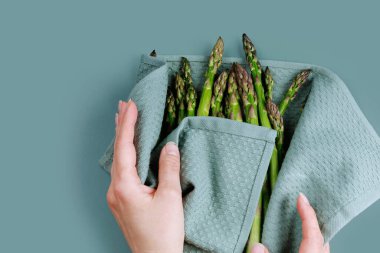 Kadınların elleri taze yeşil kuşkonmazı mutfak havlusuna sarıp hazırlanmak için mavi bir yüzeye koyuyor.