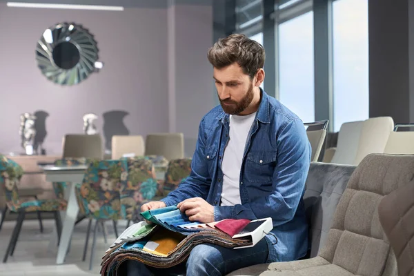 Bearded man choosing upholstery fabric color and texture from various samples in furniture store. Side view of male customer touching textile, making choise, while sitting on sofa. Concept of design.