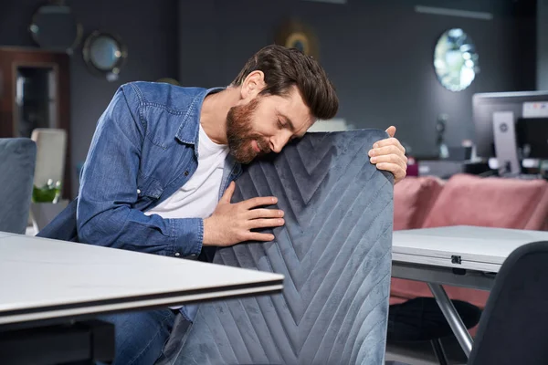 Attractive bearded man leaning to soft chair back, while exploring linear pattern before buying furniture. Handsome guy checking texture of furniture textile, while touching chair. Concept of design.