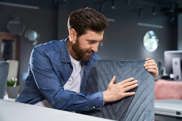 Bearded man in denim shirt exploring soft pattern chair before buying on shopping mall. Professional male designer checking texture of furniture textile, while touching chair back. Concept of design.