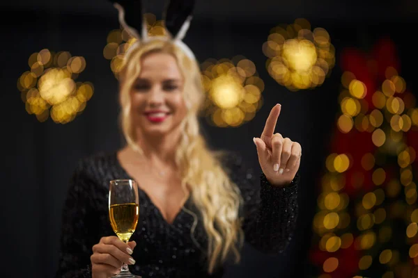 Front view of blonde, seductive woman with red lips looking at camera, smiling, holding glass of champagne. Beautiful, seductive female wearing rabbit ears. Concept of winter holiday.