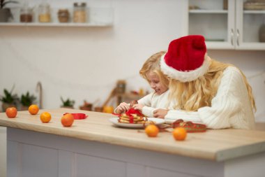 Sarışın annenin Noel şapkası takıp kızıyla masaya oturması, konuşması, gülümsemesi. Aile örgü kazak giyiyor, poz veriyor, kurabiye pişiriyor. Kış tatili kavramı.
