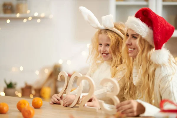 Side view of blonde woman wearing christmas hat and child wearing rabbit ears posing, smiling. Happy family having photoshoot indoors, holding decoration. Concept of winter holiday.