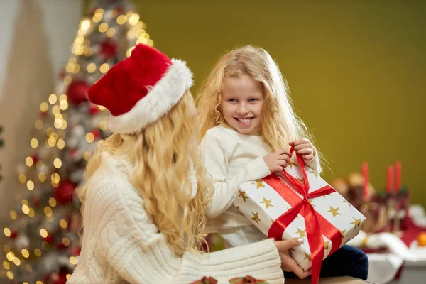 大きなクリスマスツリーで飾られたスタジオで 屋内でポーズをとって座っている幸せな家族の側面図 クリスマスの帽子の母は娘がプレゼントを持っているのを見る 家族の概念 — ストック写真